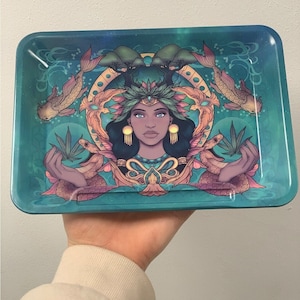 Pode incluir: Uma bandeja de rolar turquesa com uma ilustração colorida de uma mulher com uma coroa de folhas e flores. Ela está cercada por cobras, peixes e plantas. A bandeja tem um fundo brilhante, como uma galáxia.