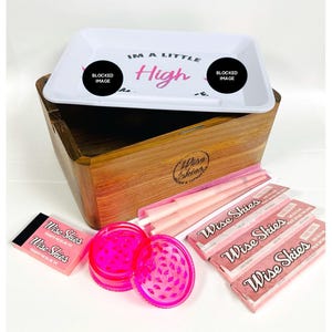Puede incluir: Una caja de madera con una bandeja de rodadura rosa y blanca, papeles de fumar rosas, un molinillo rosa y dos paquetes de papeles de fumar rosas. La bandeja de rodadura tiene el texto "I'm a little high" en ella. Los papeles de fumar est&aacute;n etiquetados como "Wise Skies".
