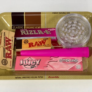 May include: A selection of smoking accessories on a gold-coloured tray. Includes a clear grinder, pink rolling papers, a pink tube, and boxes of RAW and Rizla rolling papers. The tray has text that reads "The Natural Way to Roll."