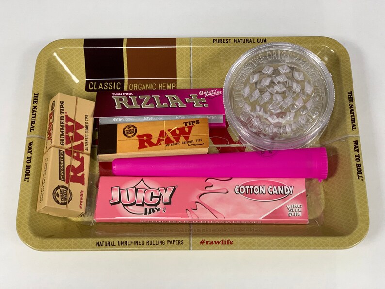 May include: A selection of smoking accessories on a gold-coloured tray. Includes a clear grinder, pink rolling papers, a pink tube, and boxes of RAW and Rizla rolling papers. The tray has text that reads "The Natural Way to Roll."