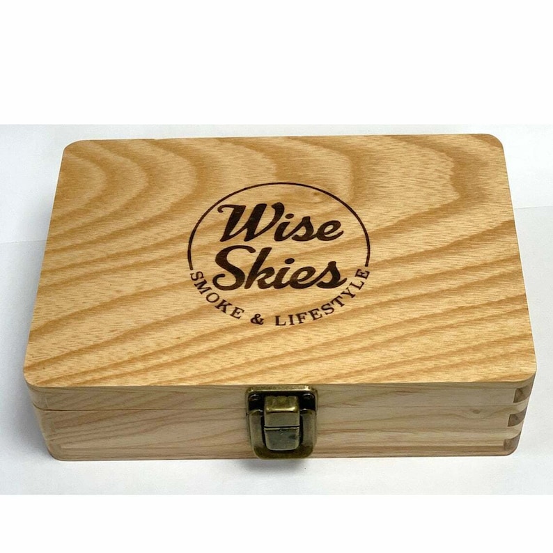 Puede incluir: Una caja de madera con un cierre de lat&oacute;n. La tapa tiene un logotipo circular con el texto "Wise Skies Smoke & Lifestyle" grabado en ella.