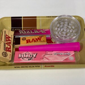May include: A rolling tray containing smoking accessories. It features a clear grinder, a pink rolling paper tube, packs of rolling papers, and a box of RAW gummed tips. The tray has text that reads "The Natural Way to Roll."