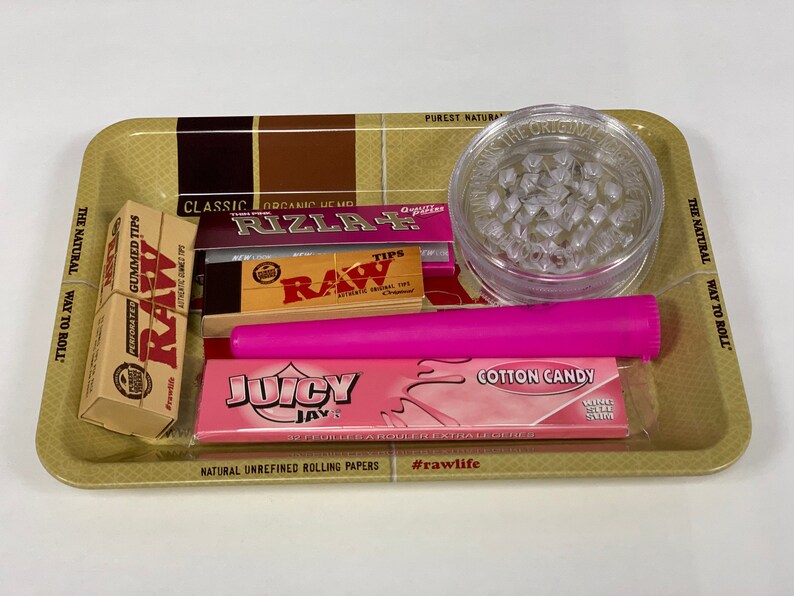 May include: A rolling tray containing smoking accessories. It features a clear grinder, a pink rolling paper tube, packs of rolling papers, and a box of RAW gummed tips. The tray has text that reads "The Natural Way to Roll."