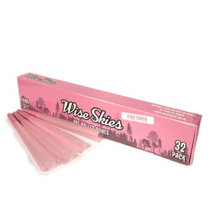 Puede incluir: Una caja rosa de conos pre-enrollados con el texto "Wise Skies" y "Pink Paper". La caja también presenta un gráfico de bosque y el texto "32 Pack". Varios conos pre-enrollados rosas son visibles frente a la caja.