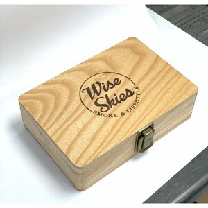 Puede incluir: Una caja de madera con cierre de pestillo. La caja est&aacute; grabada con las palabras "Wise Skies Smoke & Lifestyle" en un c&iacute;rculo.