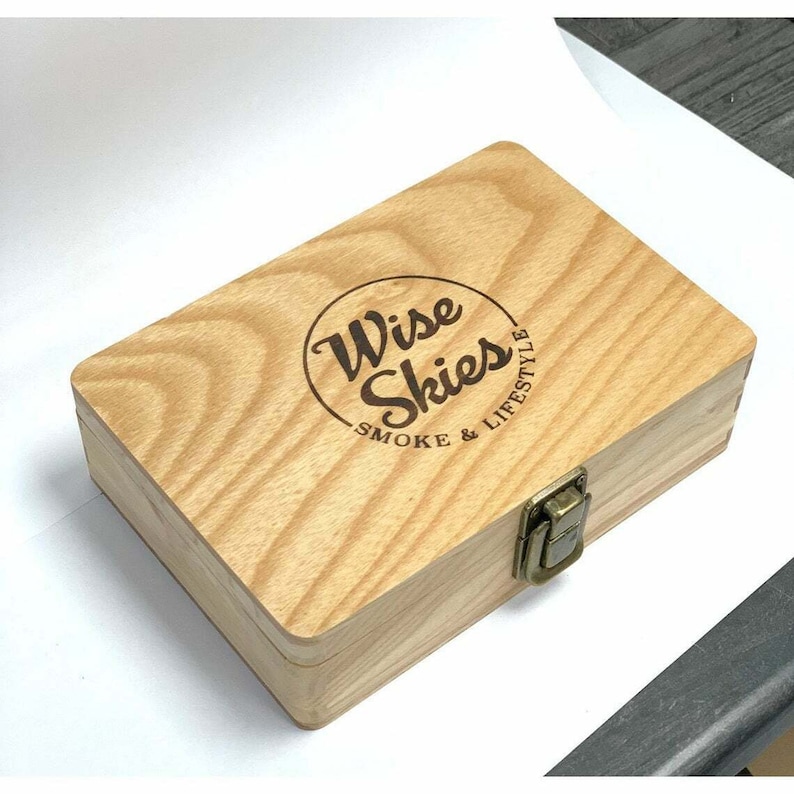 Puede incluir: Una caja de madera con cierre de pestillo. La caja est&aacute; grabada con las palabras "Wise Skies Smoke & Lifestyle" en un c&iacute;rculo.