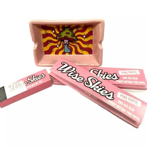 Könnte beinhalten: Rosaer Keramik-Aschenbecher mit einem Pilz- und Wirbeldesign. Zwei Packungen rosaer Drehpapiere mit dem Text "Wise Skies" und "Pink Paper King Size Slim Purest Natural Pink Paper".