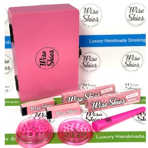 May include: Pink wooden box with a pink logo that says "Wise Skies" and the words "Smoke & Lifestyle". The box is open and contains pink rolling papers, a pink grinder, and a pink pipe. The box is on a white surface with the words "Luxury Handmade Smoking" printed on it.