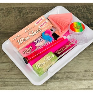 May include: A white rolling tray with various smoking accessories, including pink rolling papers, a grinder, a pink lighter, and packs of Wise Skies rolling papers.