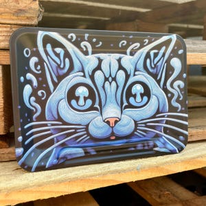 May include: A rectangular black tray featuring a blue cat face design. The cat's features are outlined in white, with mushroom-shaped pupils and a pink nose. The tray has rounded corners and a glossy finish.