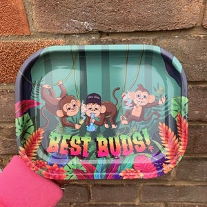 May include: A green metal rolling tray with a jungle theme featuring three cartoon monkeys and the text "Best Buds!" The monkeys are wearing hats and holding a bong.