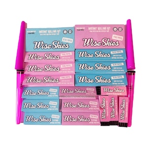 May include: An assortment of pink and blue Wise Skies rolling kits and accessories. The kits include rolling papers and tips. The text "Wise Skies" is prominently displayed on the packaging. Two pink, cylindrical accessories are on either side.