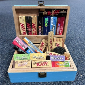 May include: A wooden box with a light blue exterior and natural wood interior, filled with various rolling papers, tips, and accessories. Brands like RAW, OCB, and Wise Skies are visible. The box is open, revealing the contents.