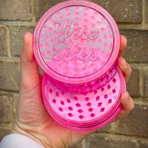 Peut inclure: Un moulin à herbe en plastique rose avec les mots "Wise Skies" gravés sur le dessus. Le moulin a un motif en diamant sur le couvercle et la base.