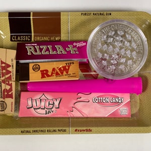 May include: A selection of smoking accessories on a gold tray. The items include a clear grinder, pink rolling papers, a pink tube, and boxes of RAW and Juicy Jay's rolling papers. The tray has text that reads "Classic Organic Hemp" and "Purest Natural Gum".