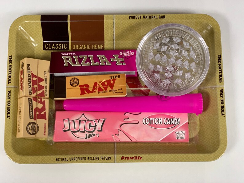 May include: A selection of smoking accessories on a gold tray. The items include a clear grinder, pink rolling papers, a pink tube, and boxes of RAW and Juicy Jay's rolling papers. The tray has text that reads "Classic Organic Hemp" and "Purest Natural Gum".