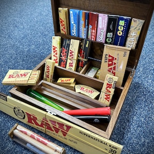 May include: A wooden box with multiple compartments filled with various brands of rolling papers and tips. The box is open and the compartments are filled with different brands of rolling papers and tips, including RAW, Elements, Skies, OCB, and ZIA. The box is made of dark wood and has a natural finish.