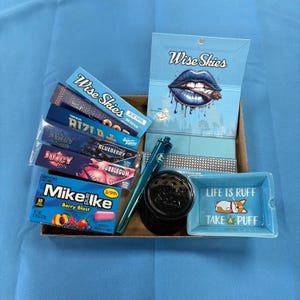 May include: A blue box filled with various smoking accessories, including rolling papers, a grinder, a pen, and a small ceramic ashtray with the text "Life is Ruff Take a Puff" and a cartoon dog. The box also contains a pack of gum.