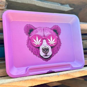May include: A rectangular rolling tray with a gradient pink and purple background. The tray features a purple bear wearing pink sunglasses with white cannabis leaf designs. The tray has a raised edge and a small lip on one side.