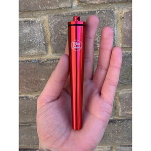May include: A red, metallic, cone-shaped container with a screw-on cap. The container has a small white logo that says "Wise Skies & Lifestyles". The container is held in a person's hand.
