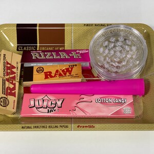 May include: A selection of smoking accessories on a gold-coloured tray. The tray includes a clear grinder, pink rolling papers, a pink tube, and various packs of rolling papers and tips. The text on the tray reads "The Natural Way to Roll."