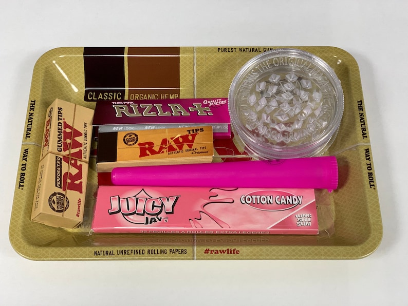 May include: A selection of smoking accessories on a gold-coloured tray. The tray includes a clear grinder, pink rolling papers, a pink tube, and various packs of rolling papers and tips. The text on the tray reads "The Natural Way to Roll."