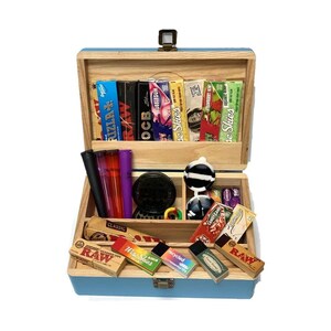 May include: A wooden box with a light blue exterior and a natural wood interior, open to reveal various smoking accessories. The box contains rolling papers, a grinder, lighters, and other items. The box is closed with a metal clasp and hinge.