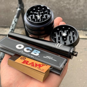 May include: A close-up of a black grinder, a black rolling tray, a pack of OCB Premium Slim rolling papers, and a pack of RAW tips. The grinder has a metal top with a circular pattern and a black base. The rolling tray is a rectangular piece of metal with a black finish. The rolling papers are in a black and white package with the text "OCB Premium Slim". The tips are in a brown and white package with the text "RAW Tips".