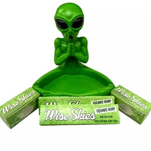 May include: Green alien figurine ashtray with a green ceramic bowl. The ashtray is surrounded by three packs of Wise Skies Organic Hemp King Size Slim rolling papers.