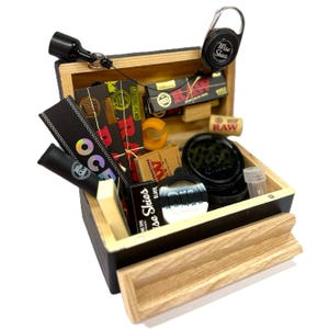 May include: A wooden box with a sliding drawer filled with various smoking accessories, including rolling papers, a grinder, a lighter, and a small container. The box is black with a wood grain pattern and has a metal latch.