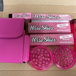 Pode incluir: Papéis de enrolar rosa e acessórios numa caixa. A caixa contém três pacotes de papéis de enrolar rosa Wise Skies, um moedor rosa, uma caixa de transporte rosa e um tubo de vidro rosa e transparente.