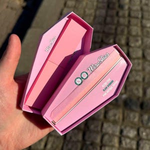 Op de afbeelding: Roze kistvormige doos met de tekst "Wise Skies" en "Ultra-Thin Slow Burning Pink Natural Papers" erop gedrukt. De doos is open en de roze rolpapieren zijn zichtbaar in de doos.