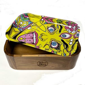 Pode incluir: Uma bandeja de metal amarela com um design psicodélico colorido que apresenta um rosto com a língua de fora e as palavras "Wise Skies" impressas na caixa de madeira.