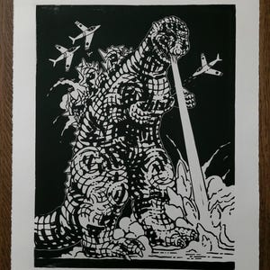 Puede incluir: Una impresión en linóleo en blanco y negro de Godzilla, un monstruo gigante, respirando fuego y destruyendo aviones de combate. La impresión está titulada "Godzilla 36" y firmada por el artista, "Michal Isaac Petrilus".