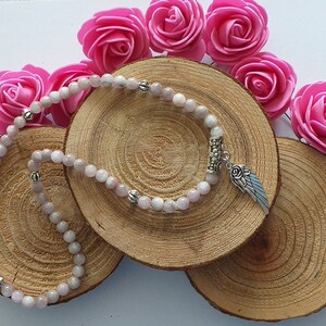 Peut inclure: Un bracelet en pierres précieuses roses et blanches avec un pendentif en forme d'aile d'ange argentée. Le bracelet est présenté sur une surface en bois avec des roses roses.