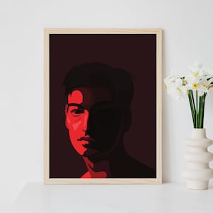 May include: A framed art print with a stylized portrait in red and black. The artwork is displayed on a white surface, with a white vase of flowers to the right. The frame is a light wood colour.