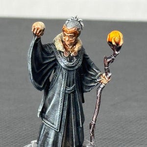 May include: A miniature figurine of a robed figure holding a skull and a glowing orb staff. The figure is on a stone base with skulls and a gold trim. The figure's robes are black, and the staff has a brown handle with a glowing orange orb.