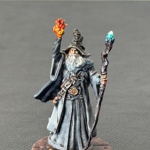 May include: Miniature wizard figurine with a long gray beard, wearing a dark gray robe and hat. The wizard holds a staff with a blue crystal and a fiery orb. The figurine stands on a round, textured base.
