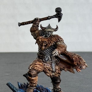 May include: A miniature figurine of a Viking warrior in a fur-lined outfit, wielding a battle axe. The figure stands atop a blue and white dragon base, with a horned helmet and detailed armor. The figurine is painted with realistic shading.