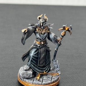 Op de afbeelding: Miniatuur fantasy figuur van een geklede tovenaar met een schedelachtig helm en staf. De figuur is geschilderd in grijstinten, zwart en goud, staand op een stenen basis met een gouden rand. De staf heeft een oranje edelsteen.