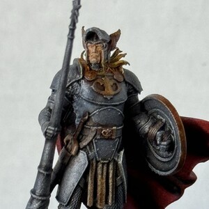 May include: A painted miniature figurine of a warrior wearing silver armor, a red cape, and a helmet with a golden crest. The warrior holds a spear in one hand and a shield in the other.