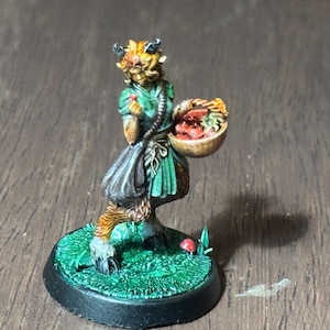May include: A painted miniature figurine of a female faun with brown horns and hooves, wearing a green and brown dress and carrying a basket of red berries. The figurine is standing on a green base with a black rim.