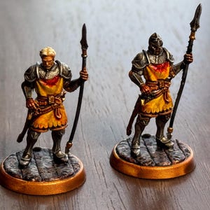 Könnte beinhalten: Zwei bemalte Miniaturfiguren von Rittern, einer mit Helm und einer mit blonden Haaren, beide halten Speere und stehen auf runden Sockeln.