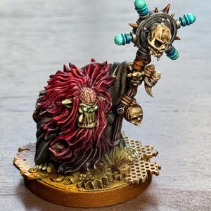 Könnte beinhalten: Detaillierte Miniaturfigur einer Fantasie-Kreatur mit rotem Haar und einem gehirnartigen Kopf. Sie hält einen Stab mit einem Totenkopf und türkisfarbenen Akzenten. Die Figur ist mit aufwendigen Details bemalt und steht auf einer strukturierten Basis mit Zahnrädern.
