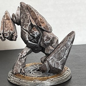 Pode incluir: Uma figura em miniatura de golem de pedra cinza com uma superf&iacute;cie detalhada e texturizada. A figura est&aacute; em p&eacute; sobre uma base circular com um design decorativo.