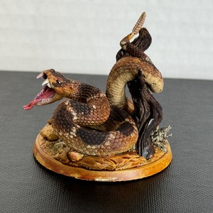 May include: A detailed figurine of a snake with an open mouth, revealing its pink tongue and white teeth. The snake is brown and black, coiled around a piece of wood. The figurine sits on a round, brown base.