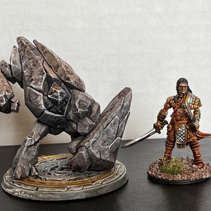 Pode incluir: Uma miniatura de golem de pedra cinza com uma textura rochosa detalhada fica em uma base circular com um design dourado e cinza. Uma miniatura pintada de um guerreiro com uma espada fica em uma base menor com detalhes marrons e verdes.