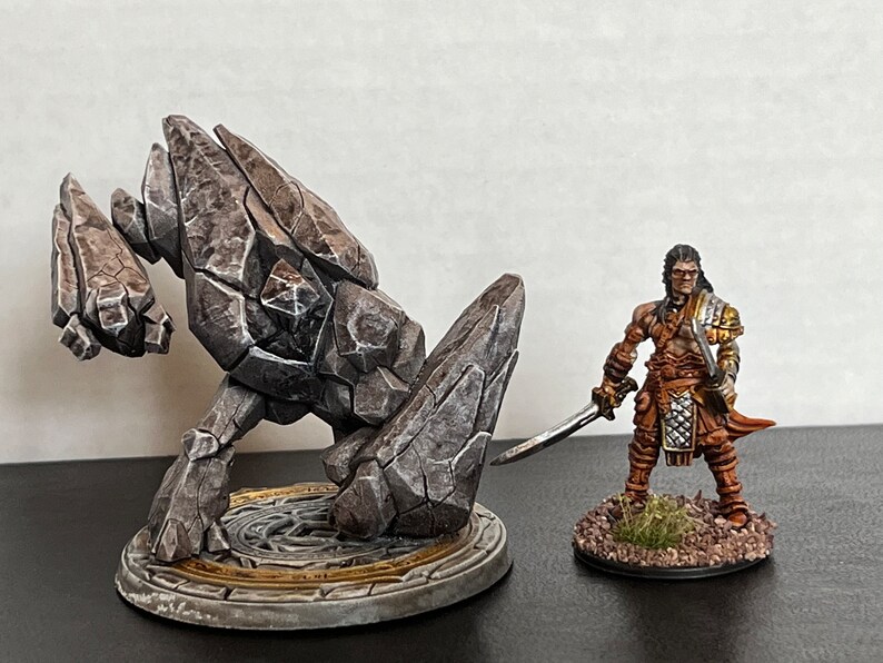 Pode incluir: Uma miniatura de golem de pedra cinza com uma textura rochosa detalhada fica em uma base circular com um design dourado e cinza. Uma miniatura pintada de um guerreiro com uma espada fica em uma base menor com detalhes marrons e verdes.