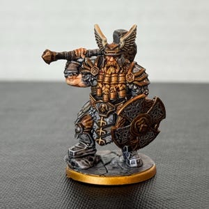 Könnte beinhalten: Miniaturfigur eines Zwergenkriegers mit Hammer und Schild. Die Figur trägt eine detaillierte Rüstung mit goldenen und bronzenen Akzenten. Der Sockel ist goldfarben, und das Farbschema ist braun, grau und bronze.