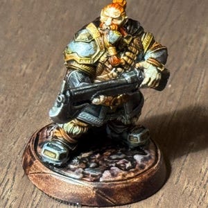 May include: A painted miniature figurine of a dwarf warrior holding a crossbow. The dwarf is wearing blue and gold armor and has a red beard. The base of the figurine is painted to look like a rocky terrain.